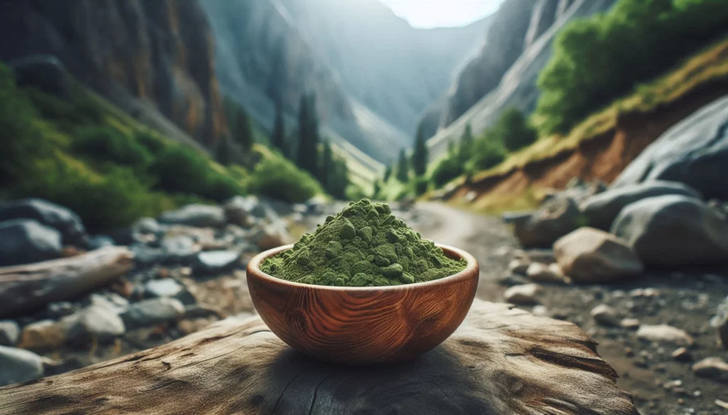 Green kratom powder in a bowl, placed on a log amidst a scenic mountain landscape with rocky terrain and lush greenery.