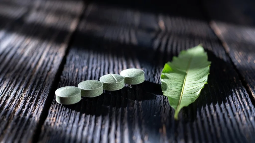 Four green 7-OH kratom tablets arranged beside a single fresh kratom leaf on dark wooden surface