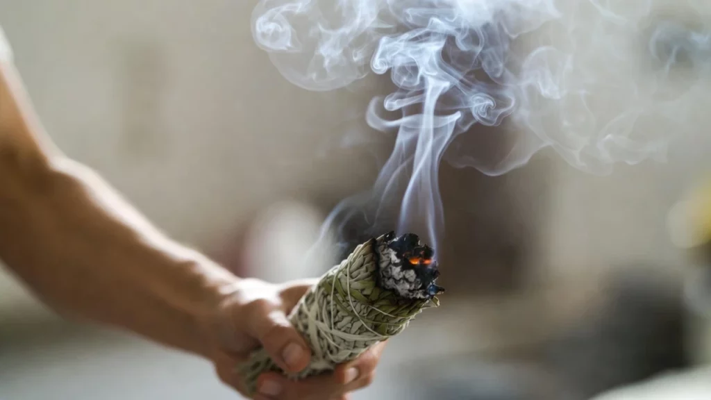 A person holding a burning white sage bundle, with smoke rising against a blurred indoor background