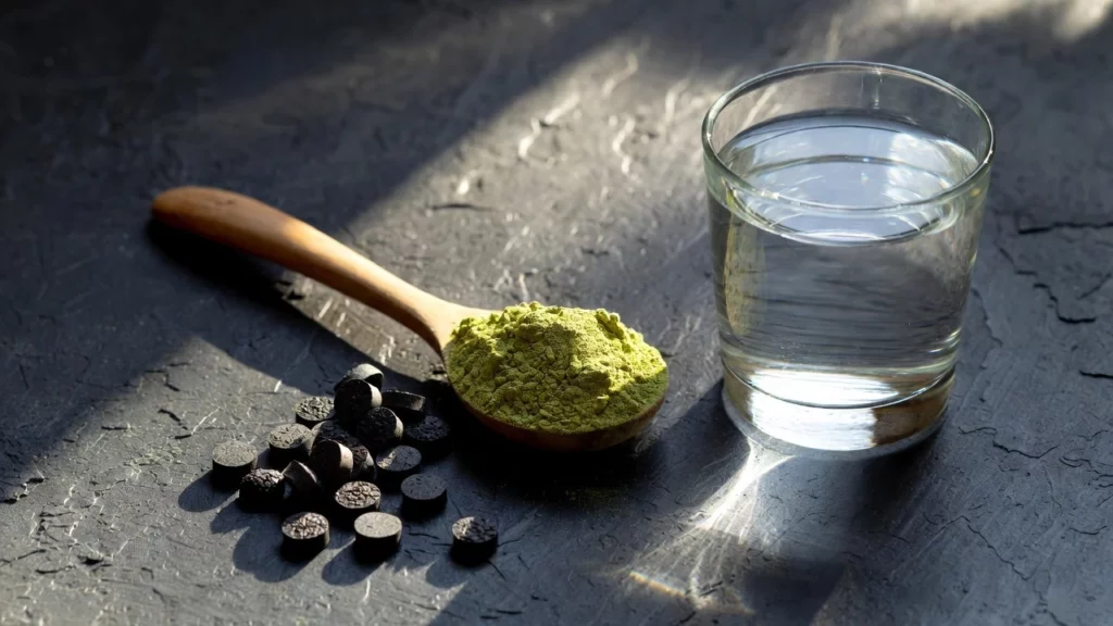 Wooden spoon holding green kratom powder next to dark shilajit tablets and glass of water on slate surface