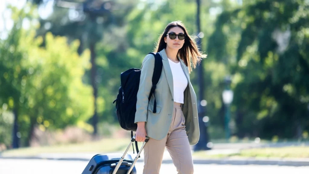A woman wearing sunglasses walks outdoors, carrying a black backpack and pulling a blue suitcase along a tree-lined path