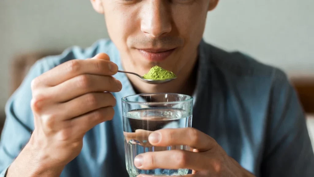 A man holding a teaspoon of kratom powder near his mouth while gripping a glass of water in the other hand