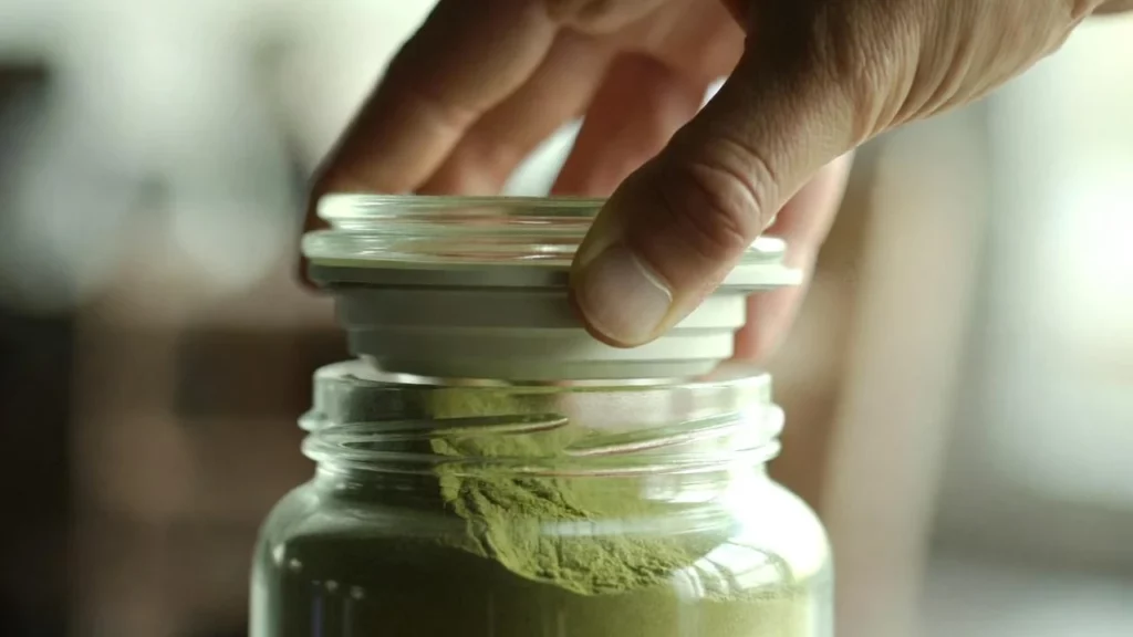 A hand placing an airtight lid on a glass jar filled with fine green kratom powder