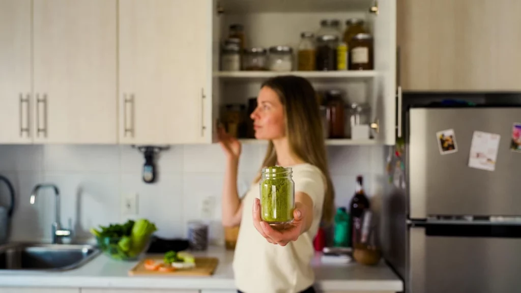 Woman in kitchen holding an open glass jar of kratom powder while standing in front of their counter