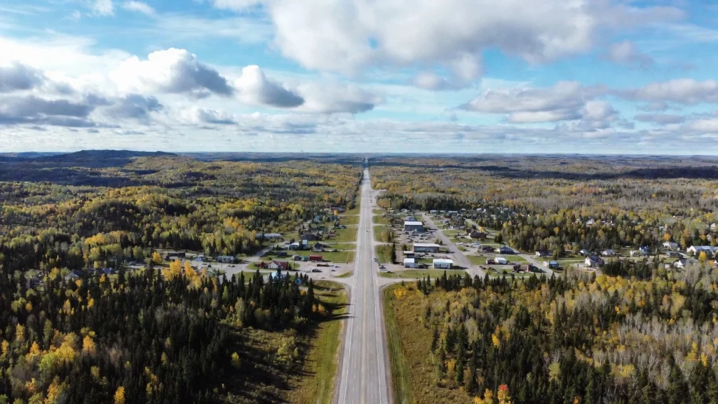 A straight rural highway cutting through dense boreal forest in Ontario during fall with yellow and orange foliage