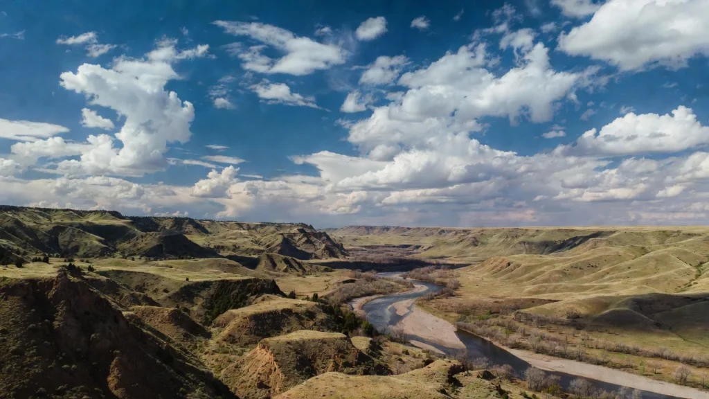 The Oldman River winding through rugged coulees and rolling prairie hills in Lethbridge, under a vast blue sky filled with scattered white clouds